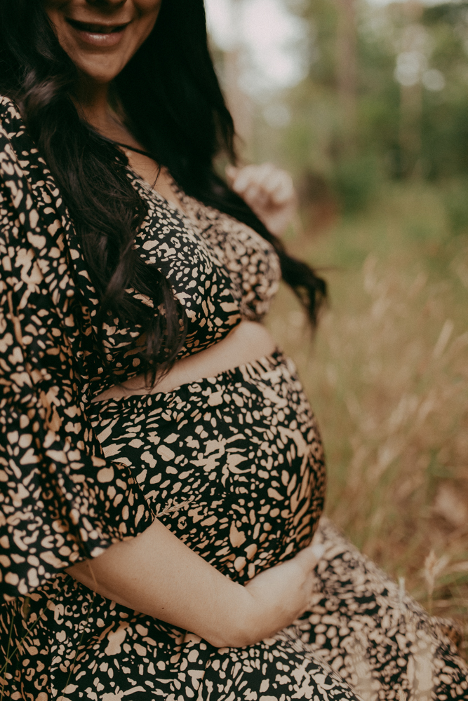 Lifestyle maternity photo of an expecting mom in a black patterned dress, softly cradling her baby bump in a natural wooded setting. Maternity photography Cary NC by Victoria Vasilyeva Photography.
