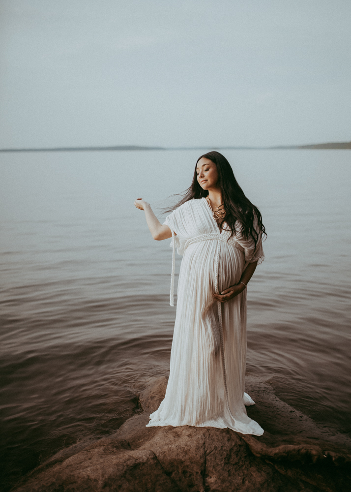 Pregnant woman with long black hair standing on a lakeside rock at sunset, wearing a soft white maternity gown and embracing her growing belly. Outdoor maternity photography Cary NC by Victoria Vasilyeva Photography.