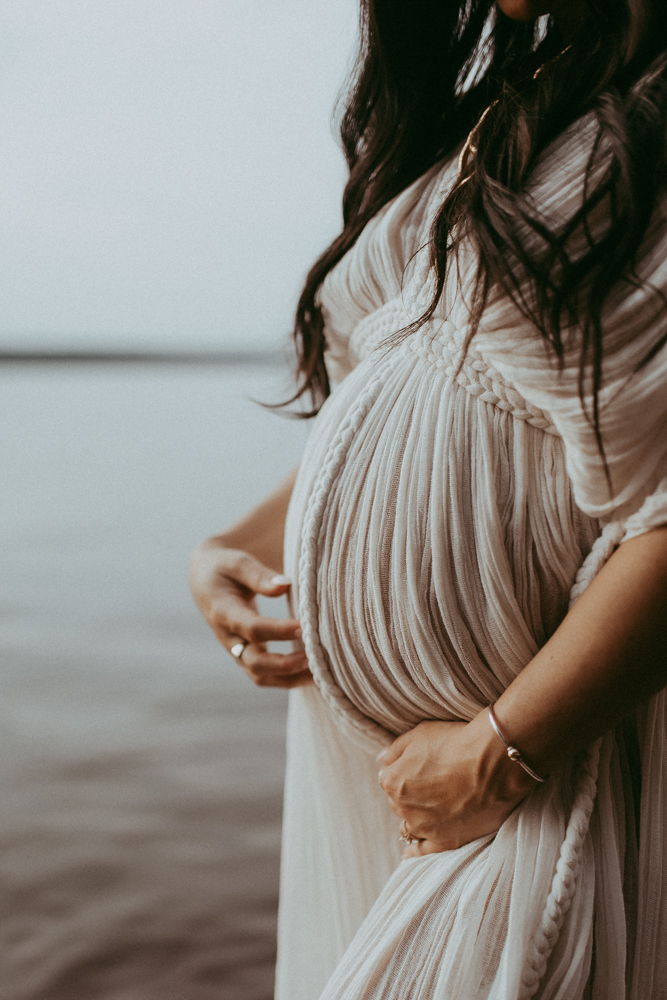 Close-up maternity portrait of an expecting mother gently holding her baby bump in a flowing neutral dress by the water.