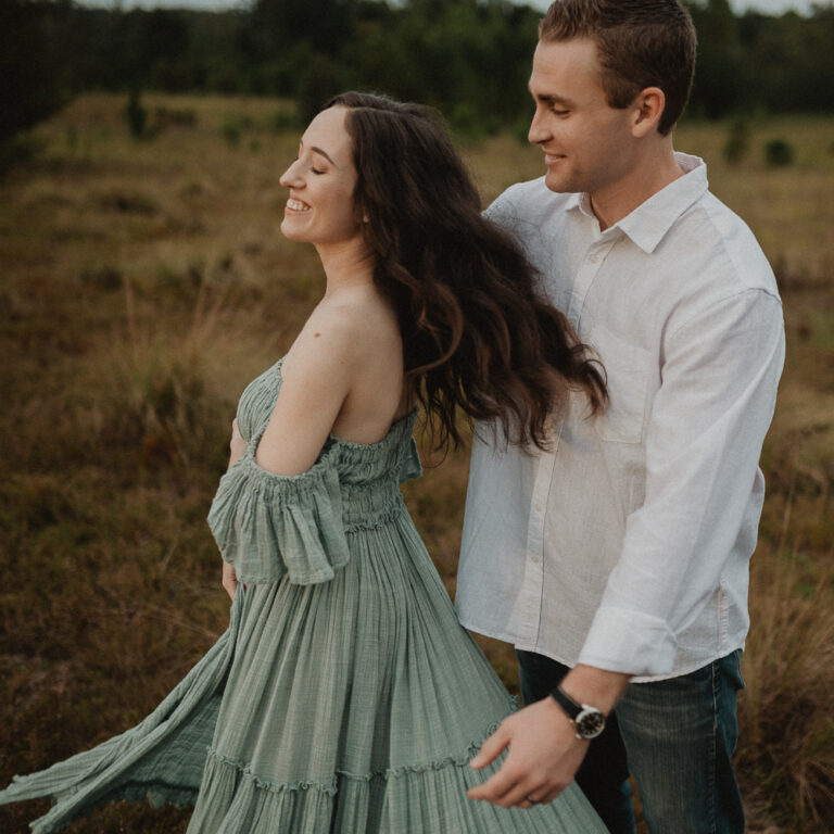 Expecting mother twirling gracefully in a flowing sage-green maternity dress while her partner watches her with a joyful smile during their outdoor sunset session. Her movement makes the dress billow softly in the breeze. Photographed by Raleigh maternity photographer Victoria Vasilyeva Photography.