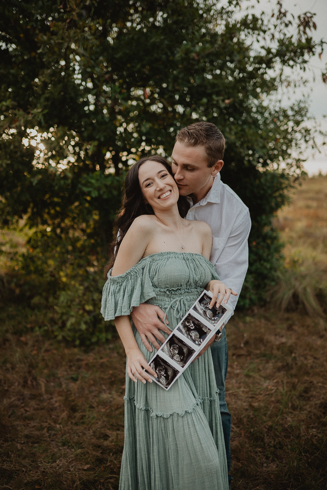 Expecting parents embracing during their outdoor maternity photoshoot, with the mother smiling brightly while holding a set of ultrasound images showing their growing baby. The father in white shirt stands behind her, wrapping his arms around her waist as sunlight glows through the background. Their journey with a Raleigh Fertility Clinic is beautifully honored in this portrait, captured by Raleigh maternity photographer Victoria Vasilyeva Photography.