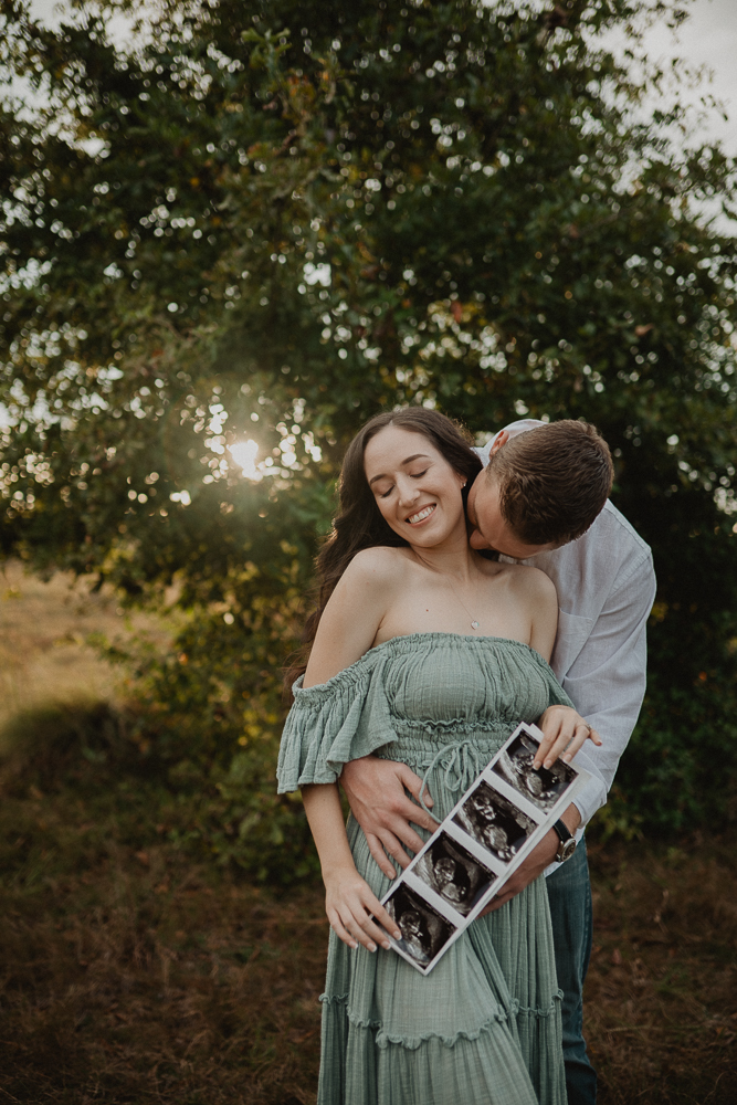 Expecting couple standing outdoors while the mother in maxi dusty blue dress holds a strip of ultrasound photos, gently leaning into her partner as golden light filters through the trees. Photographed by Raleigh maternity photographer Victoria Vasilyeva Photography.