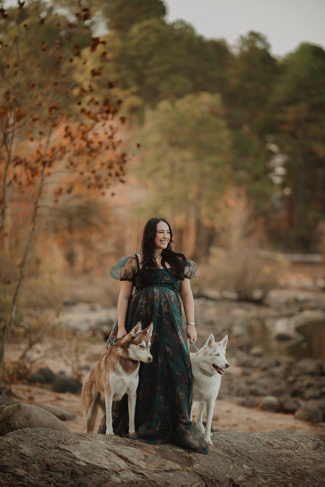 Pregnant woman in dark blue maxi dress standing on rocks with her two huskies during an outdoor maternity photo session near Greensboro, NC. Natural maternity photography by a Greensboro maternity photographer.