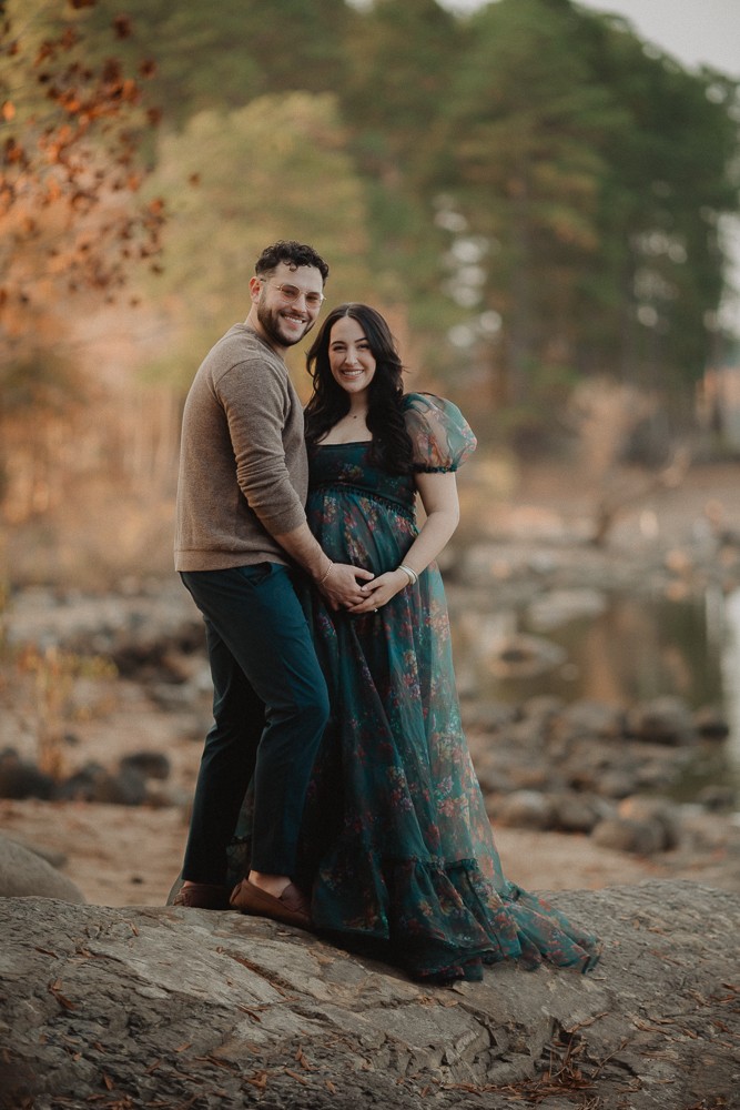 Expecting couple smiling and holding a baby bump during an outdoor maternity session by the water in Greensboro, North Carolina. Maternity photos by Victoria Vasilyeva Photography.