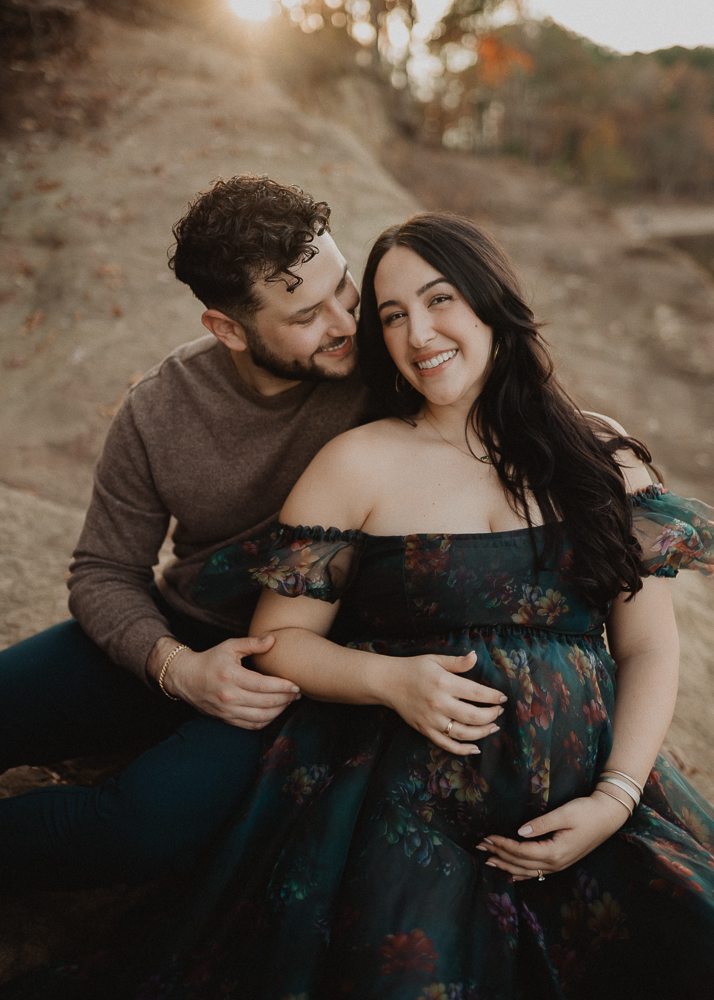 Pregnant woman with long dark hair sitting with her partner on rocks during an intimate outdoor maternity photo session in Greensboro, NC. Romantic maternity photography by a Greensboro maternity photographer.