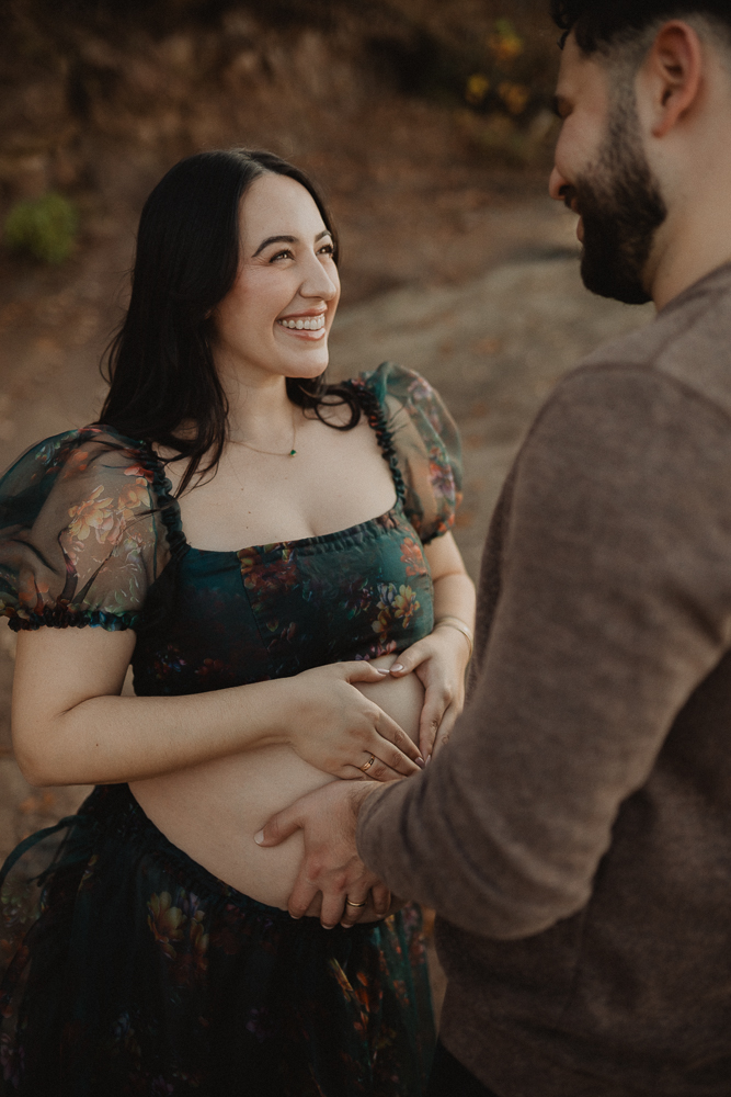Expecting mom in floral outfit smiling while her partner gently holds her bare baby bump during a maternity photo session in Greensboro, North Carolina.