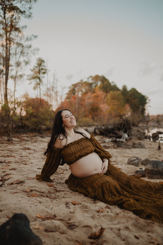 Pregnant woman sitting on sandy lakeshore during an outdoor maternity session in Greensboro, NC, wearing a brown off-the-shoulder maternity gown that highlights her baby bump. Maternity photos by a Greensboro maternity photographer.
