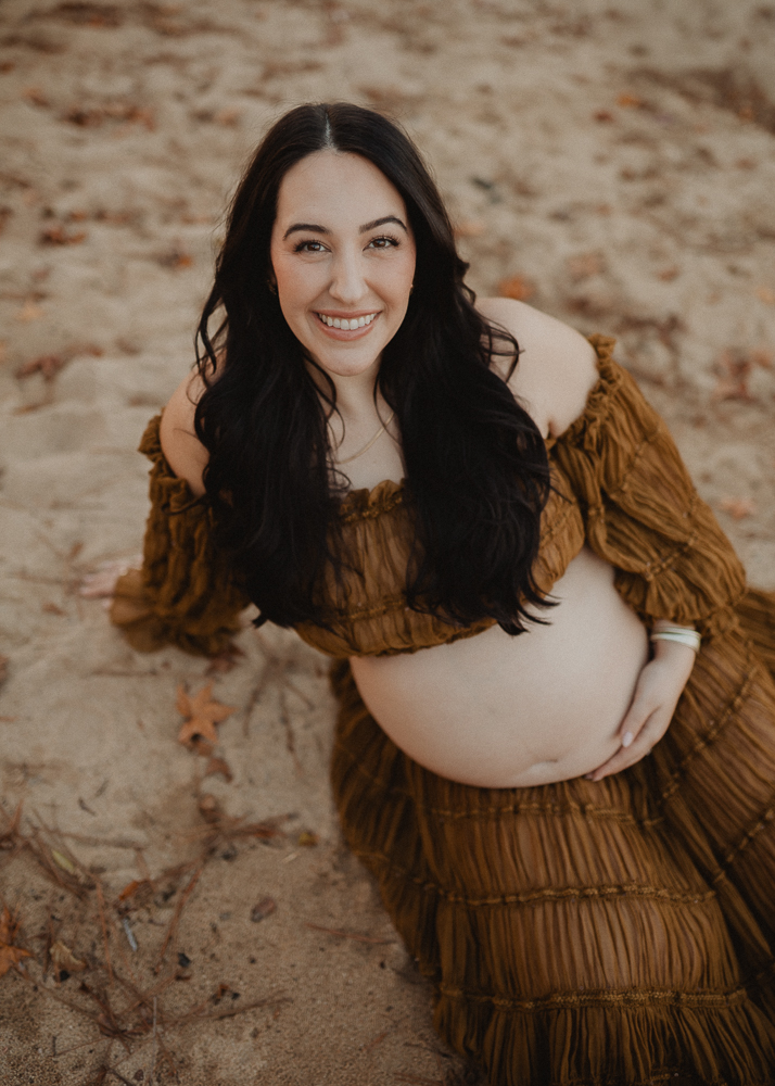 Smiling pregnant woman looking up at the camera while wearing a brown textured off-the-shoulder maternity dress during an outdoor photo session in Greensboro, NC.