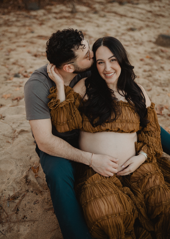 Expecting couple sitting close together on sand during a maternity session in Greensboro, NC. The mother wears a brown off-the-shoulder maternity gown, and her partner wears a short-sleeve neutral shirt and dark pants. Maternity portrait session by a Greensboro maternity photographer.