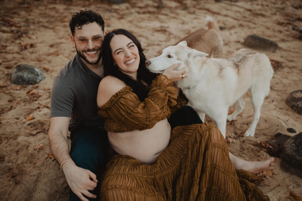 Expecting couple laughing while sitting on sand with their dogs during an outdoor maternity session in Greensboro, NC. The mother wears a rust-brown off-the-shoulder maternity dress, styled naturally by a Greensboro maternity photographer.
