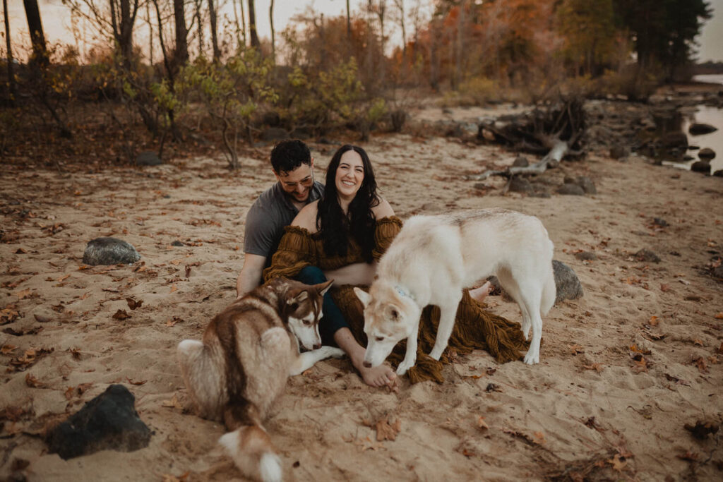 Wide maternity portrait of an expecting couple sitting on sandy ground near the lake with their dogs during a relaxed outdoor session in Greensboro, NC. The mother wears a flowing rust-brown maternity gown photographed by a Greensboro maternity photographer.