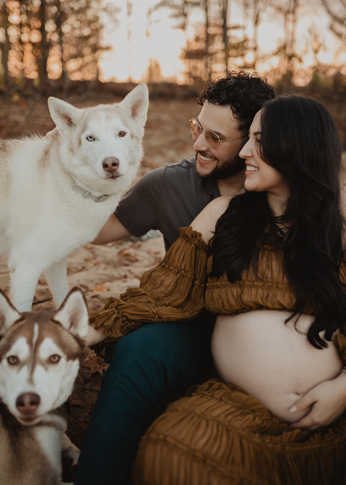 Family portrait of expecting couple seated together with their dogs in Greensboro, NC. The mother wears a brown off-the-shoulder maternity gown, styled in a natural and earthy aesthetic by Victoria Vasilyeva Photography.
