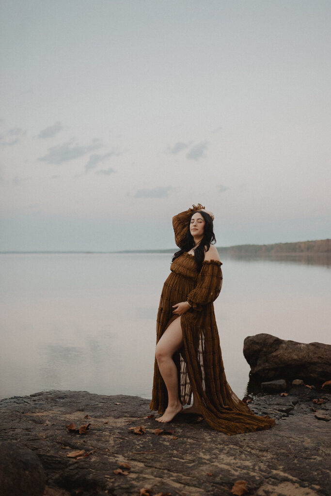 Pregnant woman standing barefoot on rocks by the lake during a maternity session in Greensboro, NC. She wears a rust-brown off-the-shoulder maternity dress with a thigh-high slit.