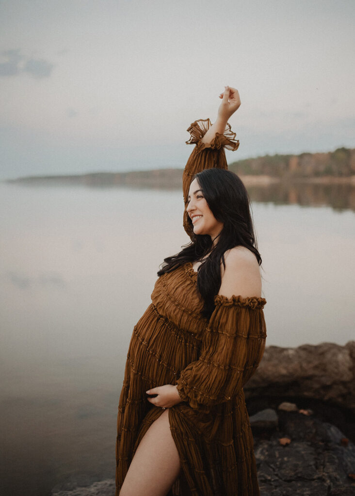Side-profile maternity portrait of a pregnant woman raising her arm while wearing a rust-brown off-the-shoulder maternity gown during an outdoor session in Greensboro, NC.