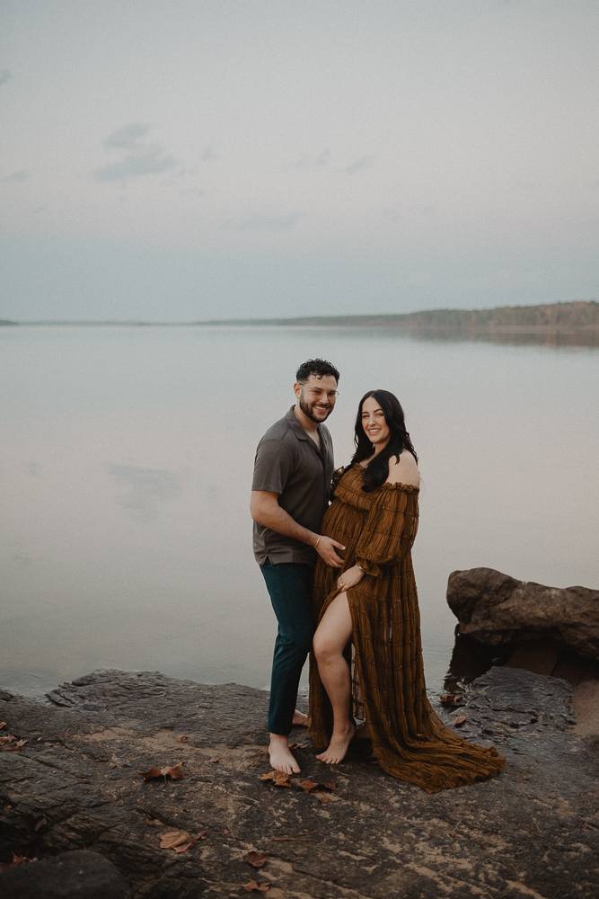 Expecting couple standing barefoot on rocky shoreline by the lake during a maternity photo session in Greensboro, NC. The pregnant woman smiles at the camera while her partner gently holds her baby bump. Maternity photography by a Greensboro maternity photographer.