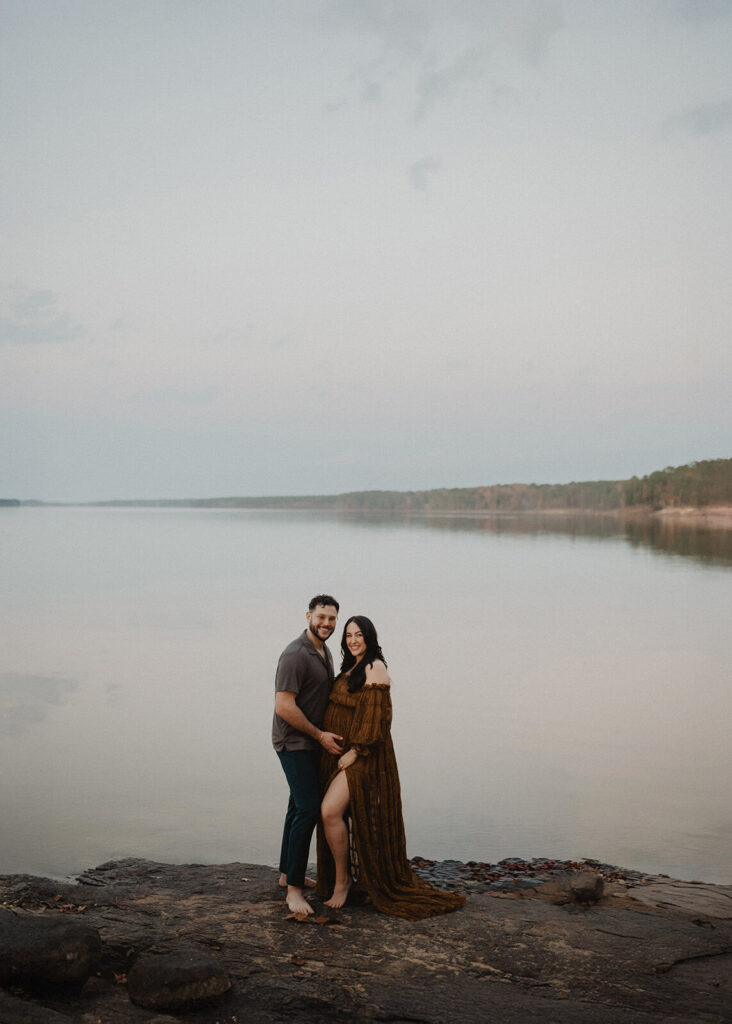 Wide maternity portrait of an expecting couple standing together by calm water in Greensboro, NC. The mother-to-be stands close to her partner as they smile softly during an outdoor maternity session with Victoria Vasilyeva Photography.