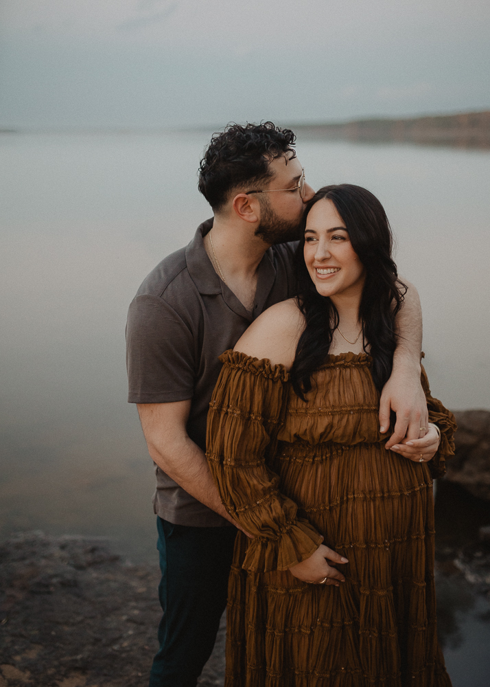Expecting father kissing the forehead of his pregnant partner while standing by the lake during a maternity session in Greensboro, NC.
