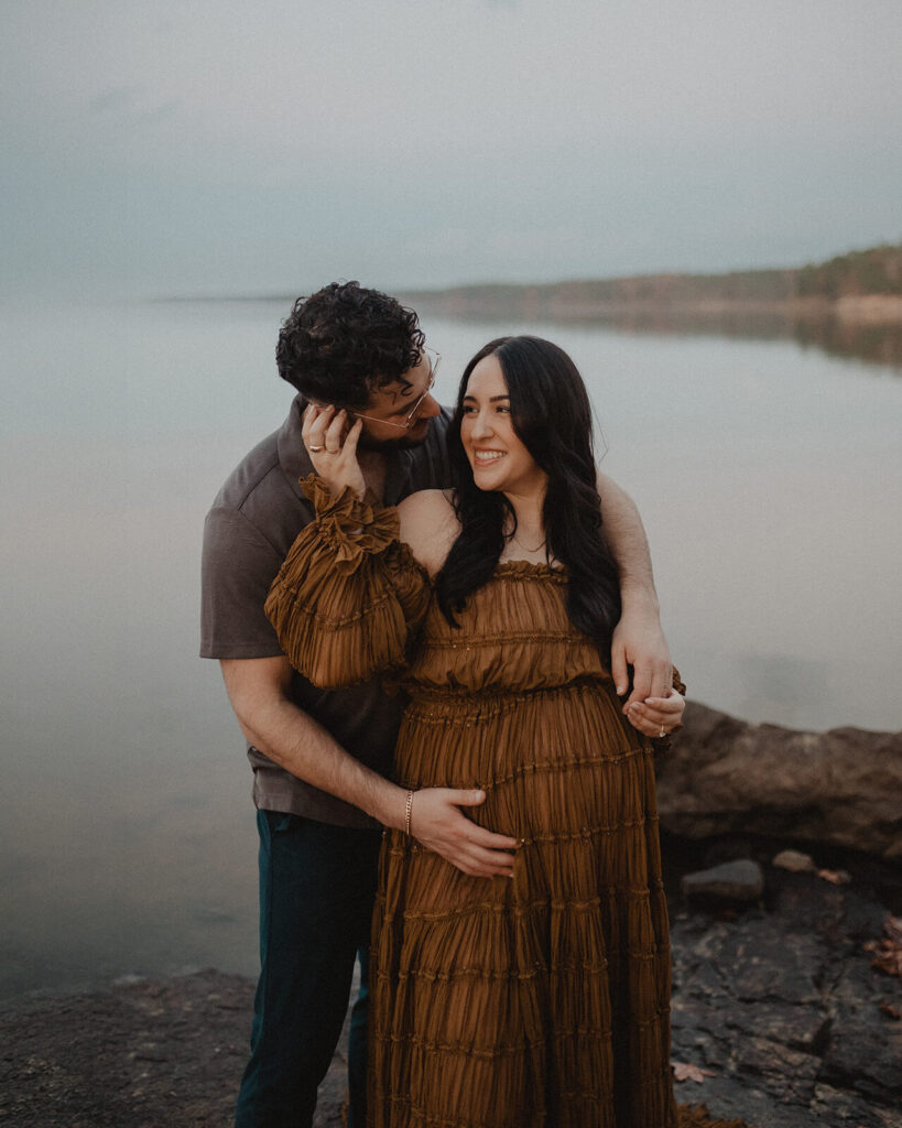 Pregnant woman smiling at her partner as he wraps his arm around her during a lakeside maternity session. Natural light photography by a Greensboro maternity photographer.