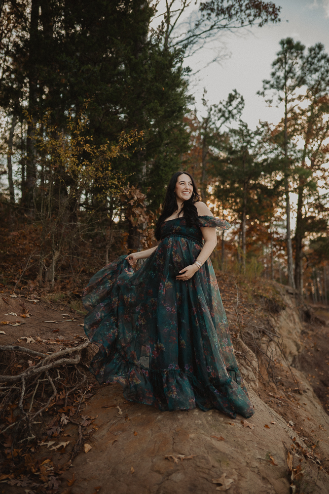 Expecting mom with dark long hair holding the flowing dark blue dress during an outdoor maternity photo session with Greensboro maternity photographer.