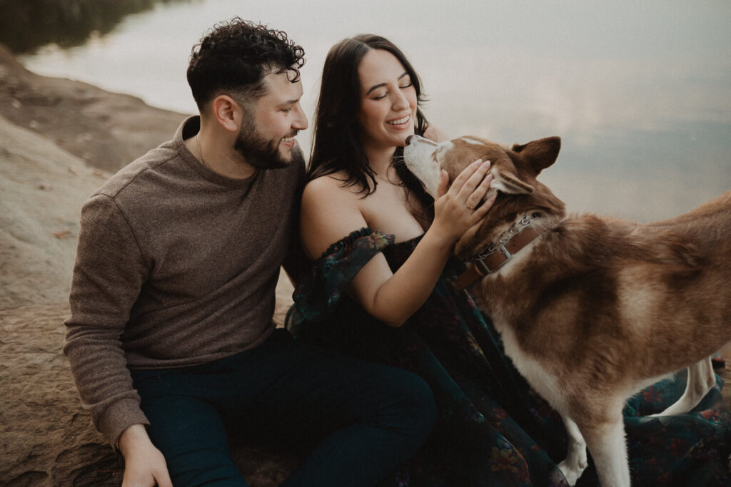 Expecting couple sitting on grey rocks by the water during a maternity session in Greensboro, NC. The mother wears a dark blue floral off-the-shoulder maternity gown, while her partner wears a neutral long-sleeve top and dark pants. Maternity photography by a Greensboro maternity photographer.