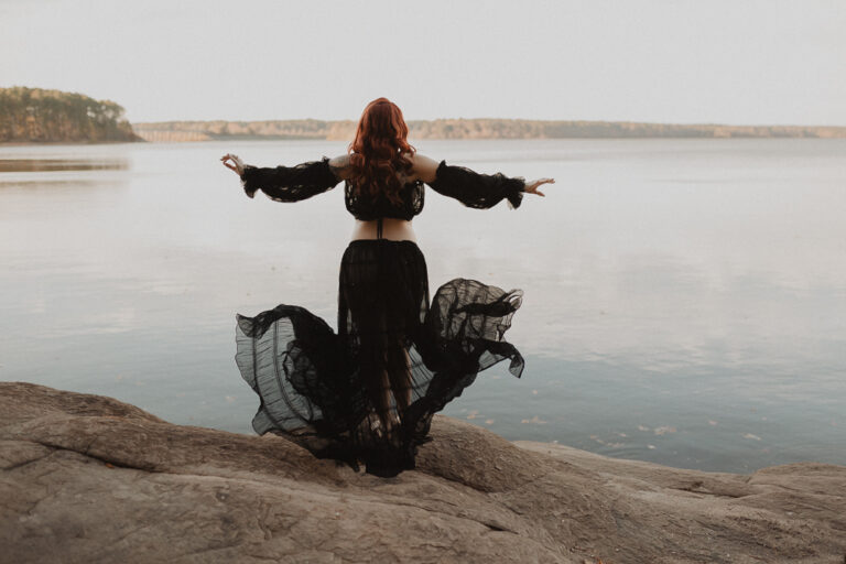Pregnant woman photographed from behind with arms extended, her sheer black maternity skirt flowing in the breeze by the lake, captured by a Fayetteville maternity photographer - Victoria Vasilyeva Photographer.