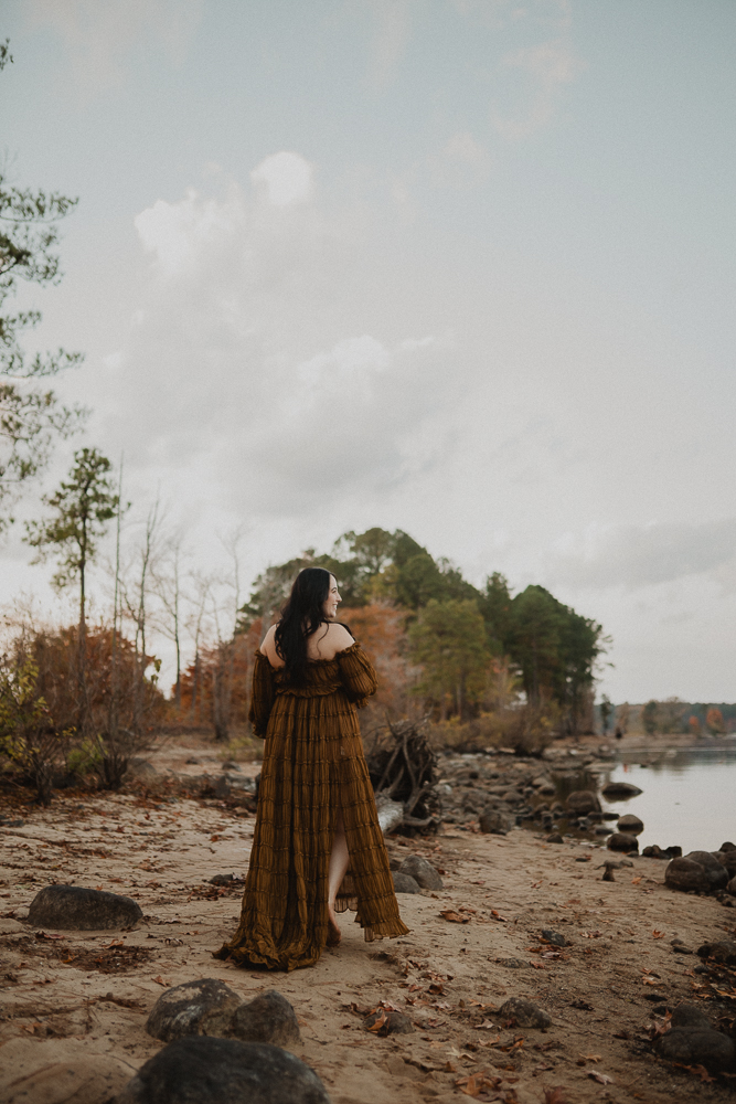 Pregnant woman walking barefoot along a sandy shoreline during an outdoor maternity session in Greensboro, NC. She looks toward the lake.