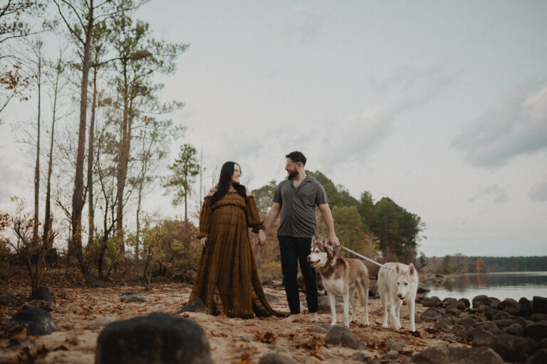 Expecting couple walking together with their dogs along a sandy lakeshore during a maternity session in Greensboro, NC. Maternity photo session with dogs.
