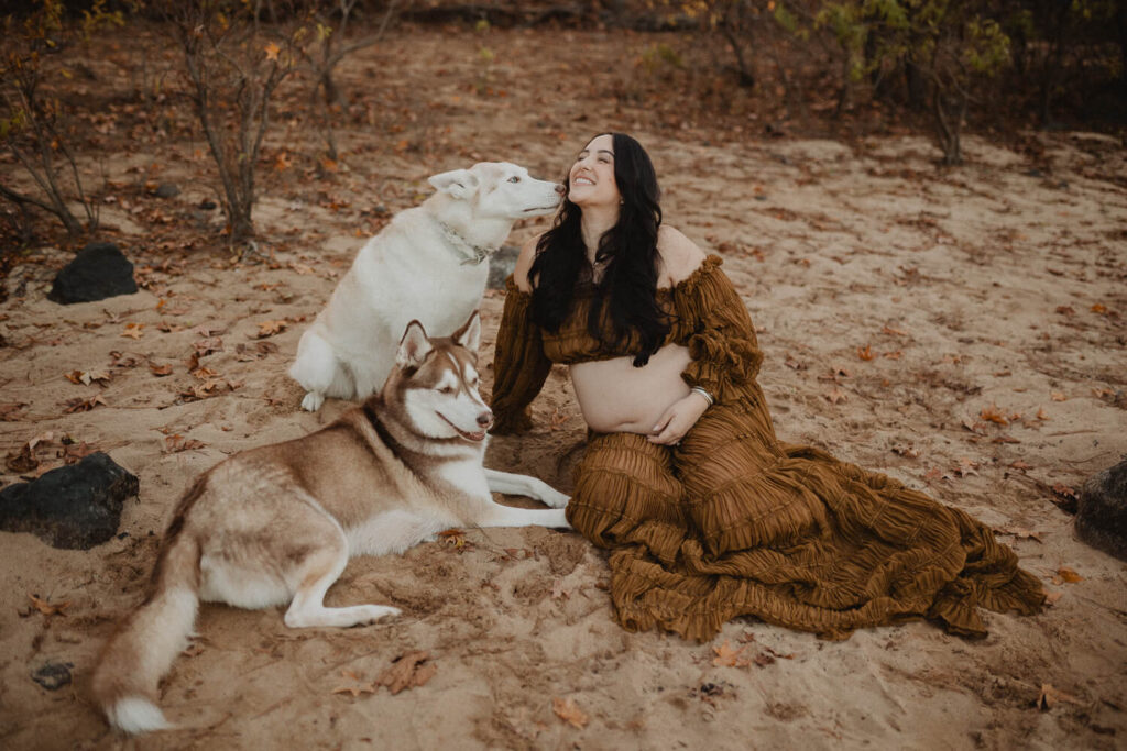 Pregnant woman sitting on sand near the lake surrounded by her dogs during a relaxed maternity session in Greensboro, NC.