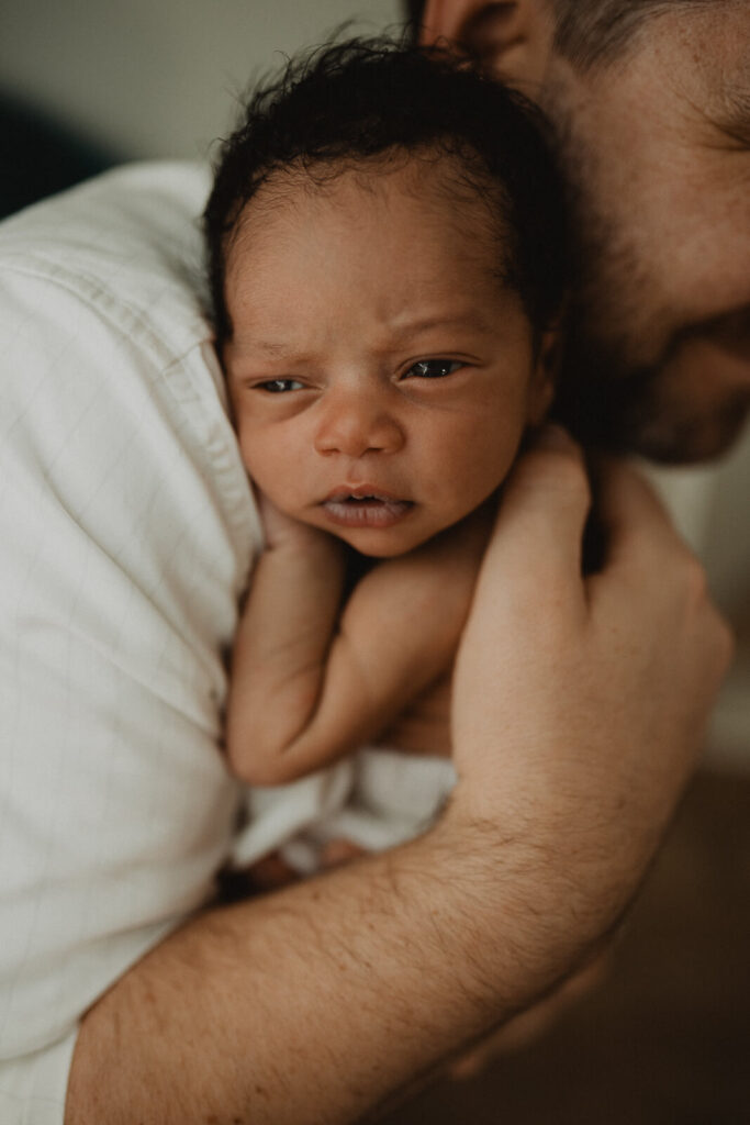 A wide-eyed newborn baby boy rests safely against dad's shoulder during a cozy lifestyle newborn session photographed by Raleigh newborn photographer Victoria Vasilyeva Photography, documenting authentic family connections at home.