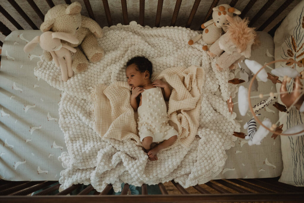 Overhead view of a newborn lying in a beautifully styled crib surrounded by soft blankets, heirloom toys, and a nursery mobile, photographed by Raleigh newborn photographer Victoria Vasilyeva Photography.