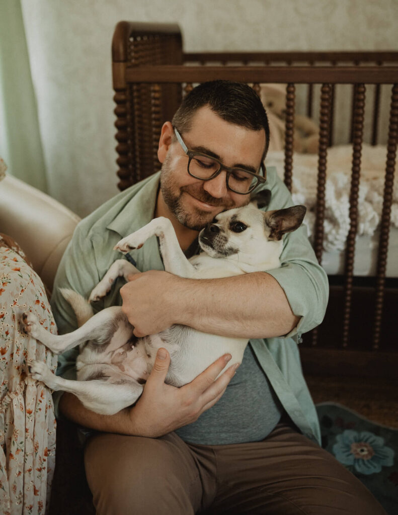 Goodnight Sweet Baby. Dad cuddles the family dog beside the nursery crib during an in-home newborn session photographed by Raleigh newborn photographer Victoria Vasilyeva Photography, highlighting the importance of including beloved pets in family memories.