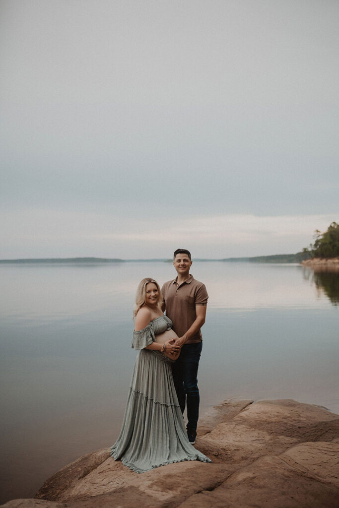 Expecting couple standing by calm lake waters during a maternity session with a Wake Forest photographer.