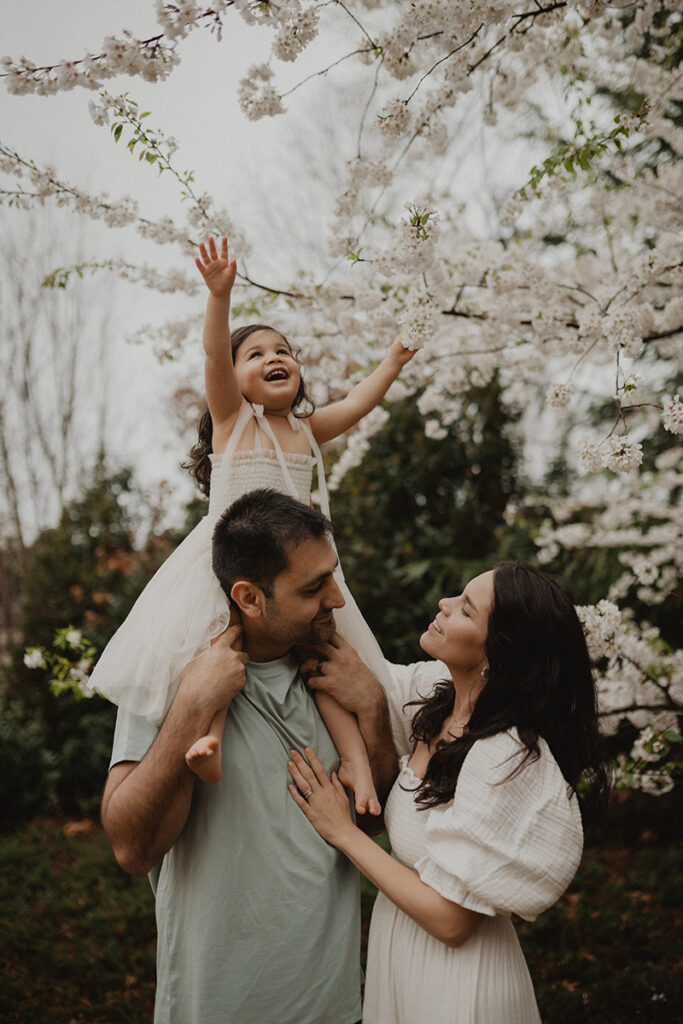 Natural outdoor family portrait under flowering trees in Chapel Hill, North Carolina, photographed by Victoria Vasilyeva Photography, featuring parents smiling at their daughter during a spring family photo session.