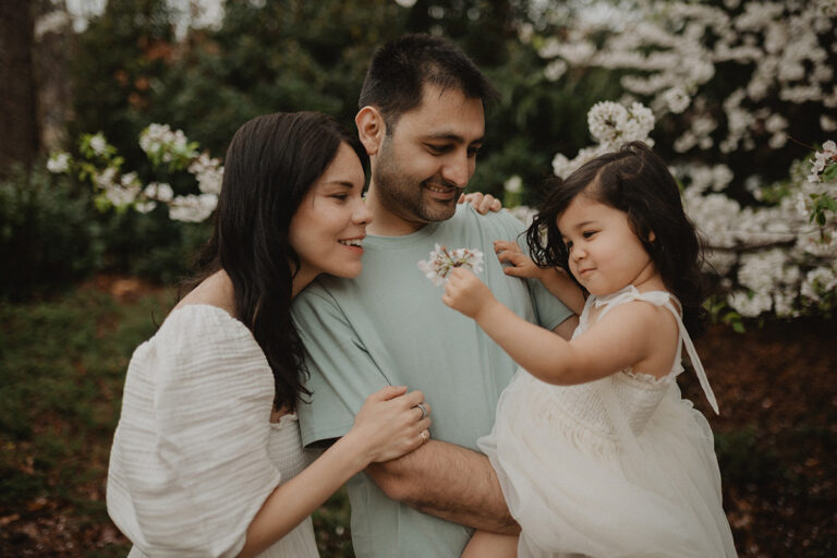 Victoria Vasilyeva Photography documenting a sweet family moment as a young girl admires cherry blossoms while held by her parents in a blooming garden in Chapel Hill.