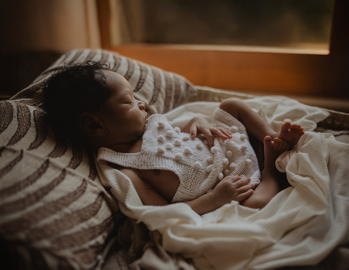 A peaceful newborn baby boy sleeps by the window wrapped in soft neutral blankets and a textured knit romper during an in-home session captured by Raleigh newborn photographer Victoria Vasilyeva Photography, celebrating the beauty of early parenthood.
