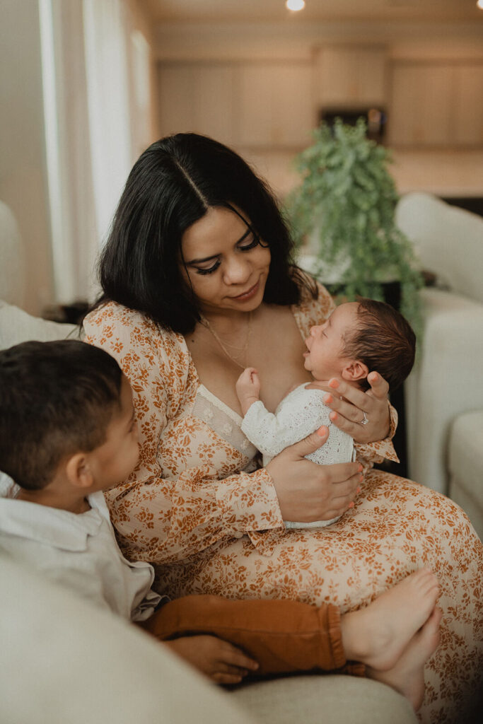 Mother holding her newborn while sitting with her toddler during a relaxed in-home family session, near Honeysuckle Lactation Consultants in Holly Springs, NC.