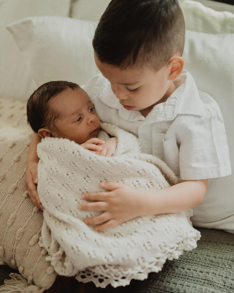 Older sibling gently holding a newborn brother wrapped in a knit blanket during newborn session with Raleigh newborn photographer.