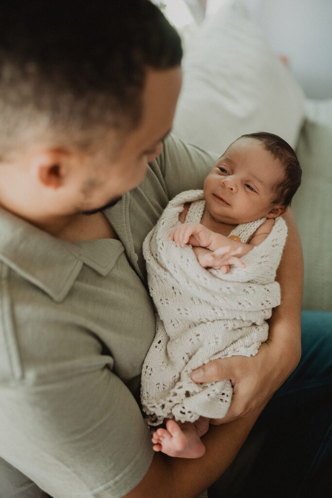 Father cradling an awake newborn baby boy wrapped in a soft knit blanket during an in-home lifestyle session.