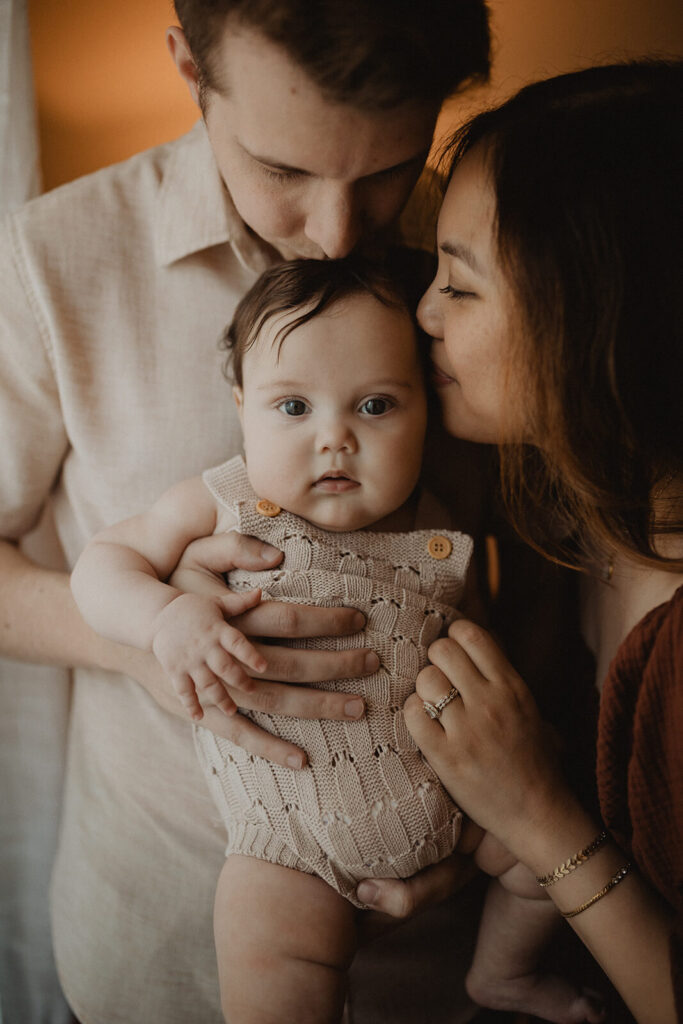 Mother and father cuddling their 5 months old baby boy and sharing affectionate kisses during an in-home family session, captured by Raleigh family photographer.