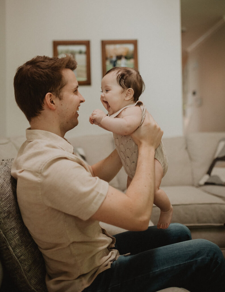 Father holding his laughing baby boy during a playful moment in the living room, photographed by Victoria Vasilyeva Photography.