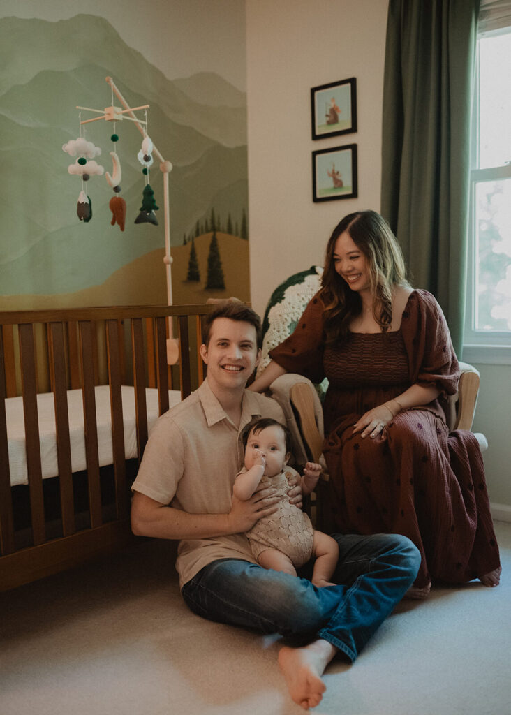 Parents and baby sitting together in a beautifully designed nursery with mountain mural and crib, photographed by Raleigh family and newborn photographer.