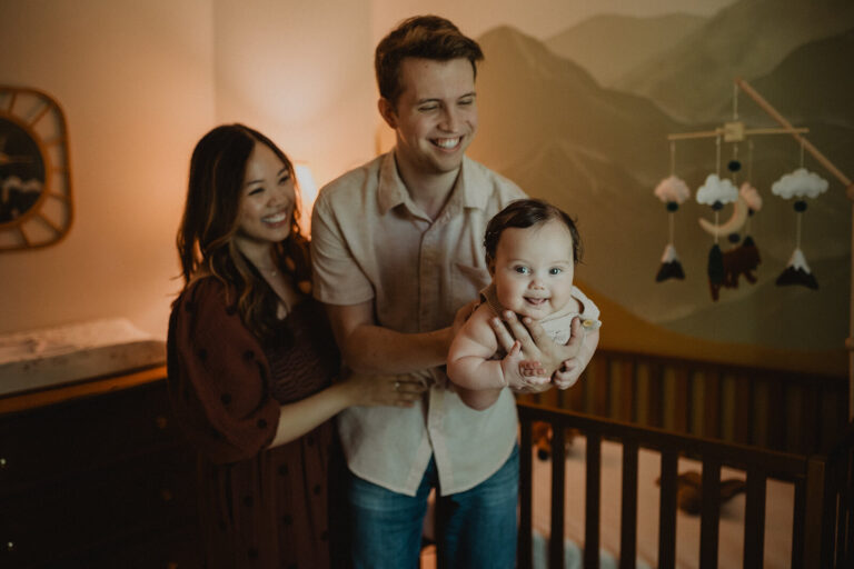 Smiling baby boy held by parents beside the crib during an in-home family photography session connected to a Raleigh sleep coach interview.