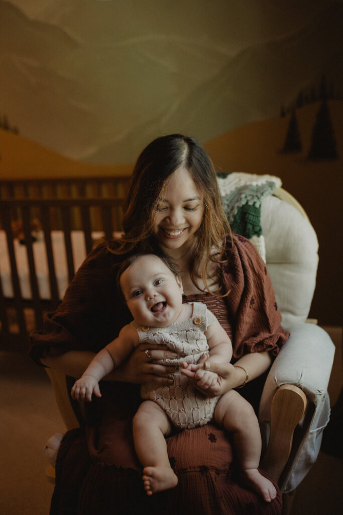 Mother in brown dress holding her happy baby boy in a nursery rocking chair during a calm bonding moment.
