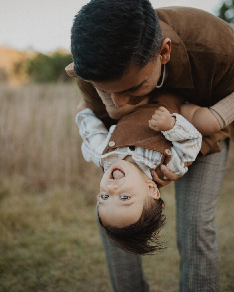 Father playfully holding his laughing baby boy during a candid pumpkin patch fall family photo session at Hill Ridge Farms in Youngsville, NC.