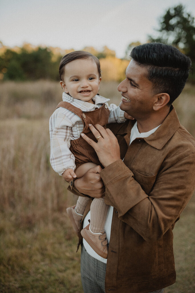 Father holding his smiling son during an outdoor fall family photography session at Naylor Family Farm & Corn Maze in Fuquay-Varina, NC. Family photo session by Victoria Vasilyeva Photography.