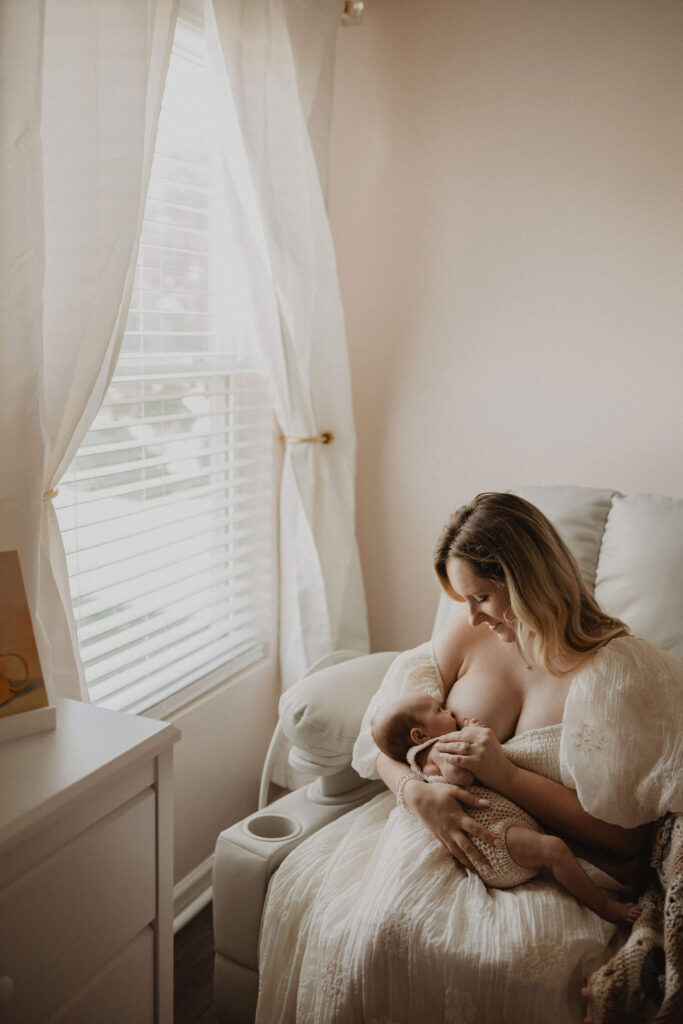 Mother in white dress breastfeeding her newborn baby girl by a bright nursery window during a natural in-home newborn photography session with Victoria Vasilyeva Photography in Raleigh NC.