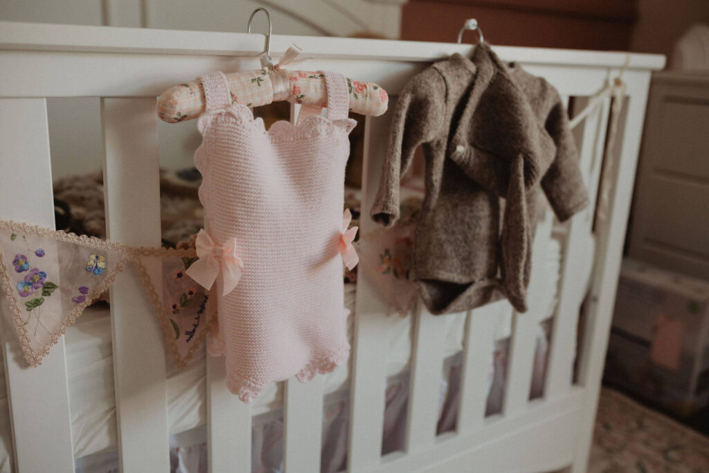 Pink knitted baby romper and neutral newborn outfit hanging on a white crib in a baby girl's nursery, photographed by Victoria Vasilyeva Photography in Raleigh NC.