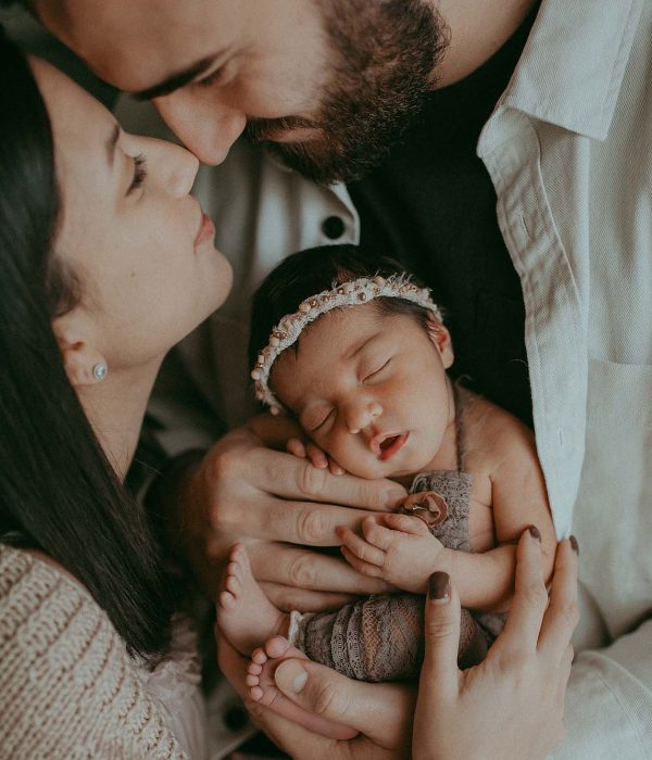 tiny newborn baby girl in brown lace romper is in dad's hands. He holds her in front of camera. Mom hugs them both and looks up to dad. Newborn photo session by Victoria Vasilyeva Photography - Fayetteville NC newborn photographer.
