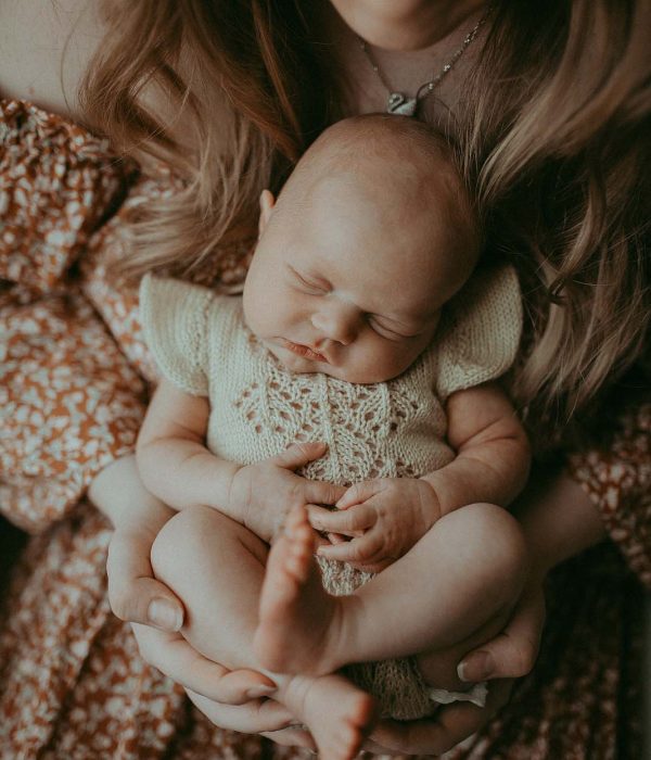 Newborn Baby girl is sleeping in her mom's lap. Mom holds her baby girl with 2 hands and looks down at her. The newborn portrait was taken during in-home newborn session with Chapel Hill Newborn Photographer - Victoria Vasilyeva Photography.