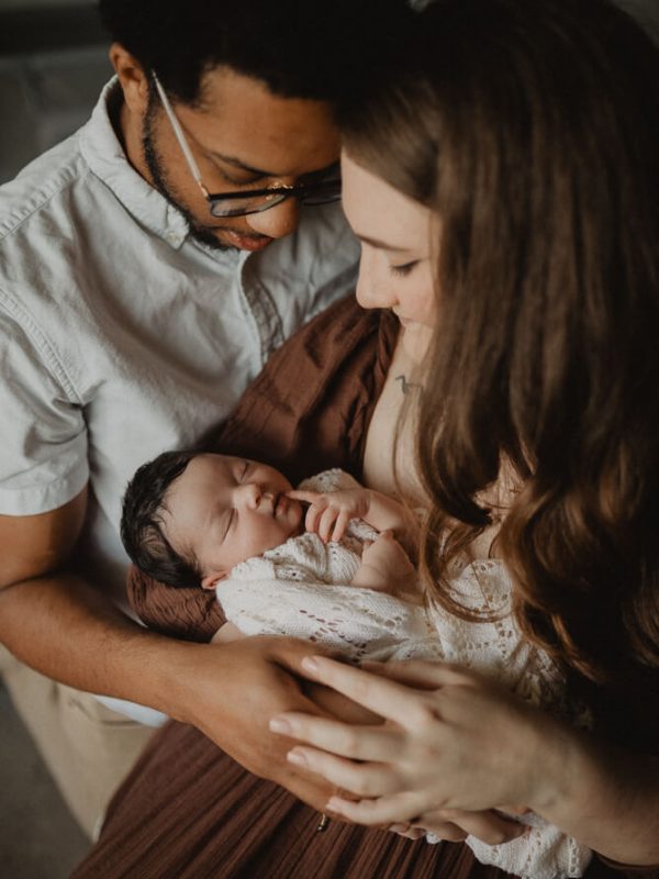 New parents cuddling their newborn during an in-home newborn photo session in Cary, NC. The mother in a brown dress and father in a white shirt lean their foreheads together while holding their baby on a cozy bedroom bed. Captured by Victoria Vasilyeva Photography - Cary newborn photographer.